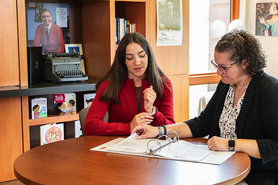 Two women engaged in a discussion at a table, reviewing documents while sitting in a cozy library setting.